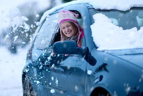 Smiling woman in wooly hat looks out of her car window. Snowy scenery.